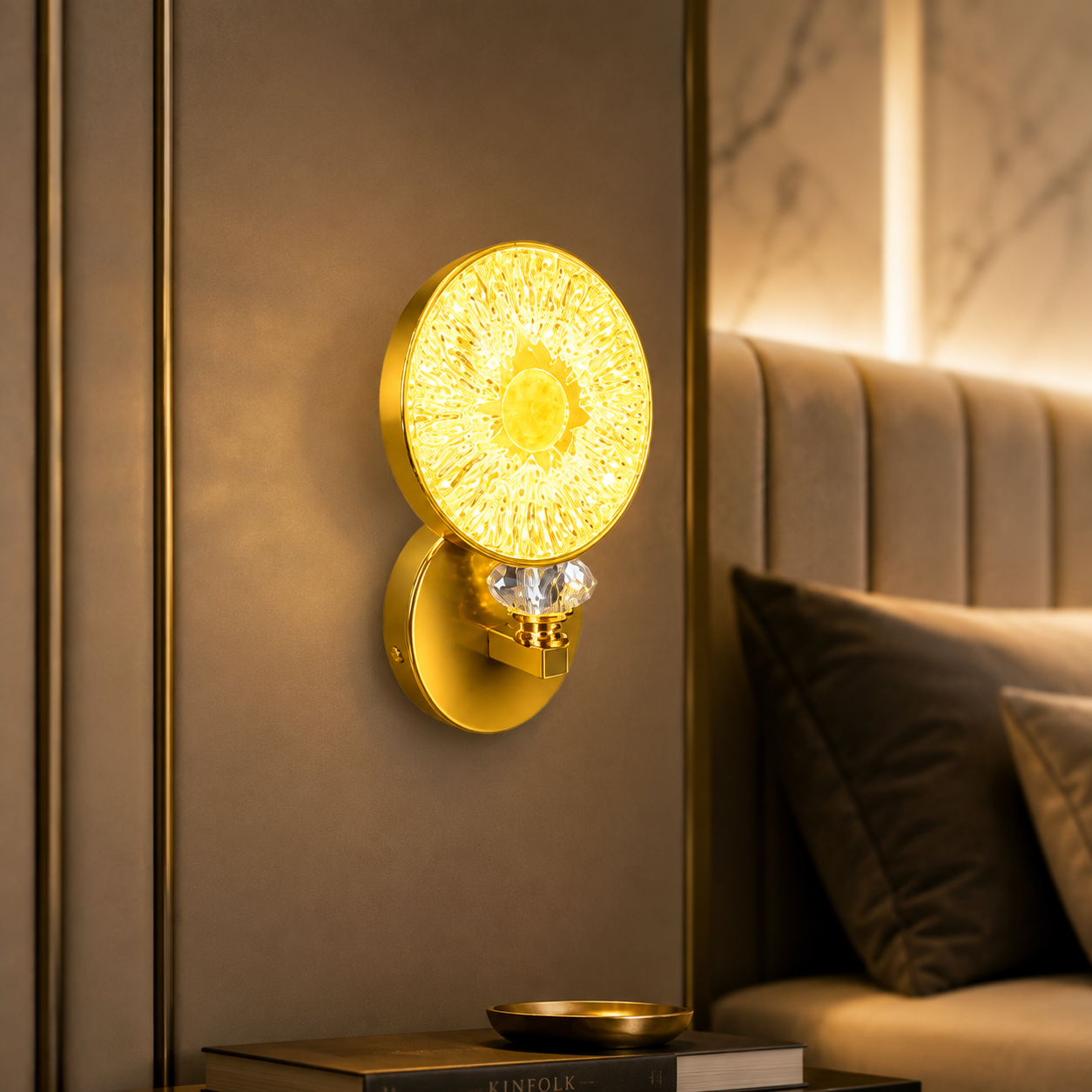 A modern wall-mounted light fixture in a warm gold finish, featuring a circular textured shade and a faceted crystal accent, shown lit in a luxurious bedroom setting.