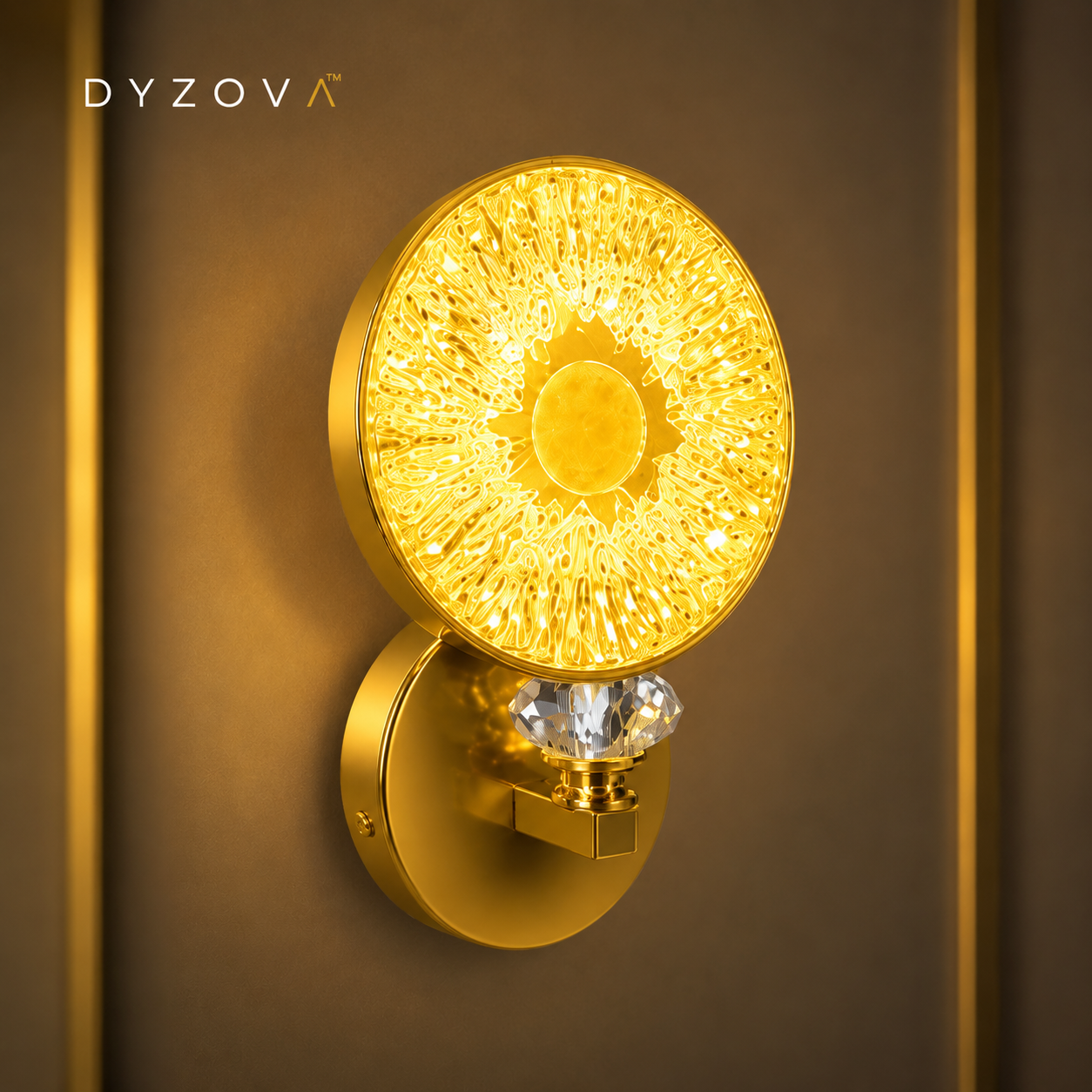 A modern wall sconce light fixture with a circular gold-toned face, a faceted clear crystal accent, and a matching round wall mount, shown lit against a warm brown background. The brand name DYZOVA is visible in the top left corner.