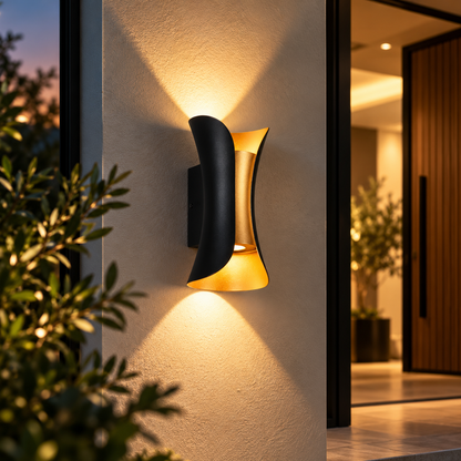 A modern curved wall sconce light fixture in matte black and warm gold, mounted on a textured beige wall and illuminated at dusk, with blurred greenery in the foreground and a doorway in the background.