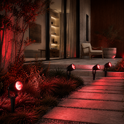 Outdoor landscape lighting system with multiple black ground spotlights casting a warm red glow over a stone pathway, plants, and a modern patio area at night.