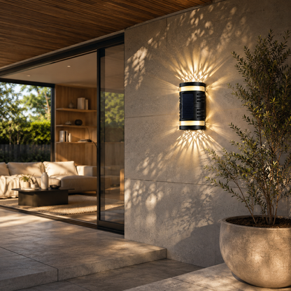 A modern cylindrical outdoor wall light with a black body and gold trim, emitting a starburst of warm light onto a stone wall next to a potted plant.