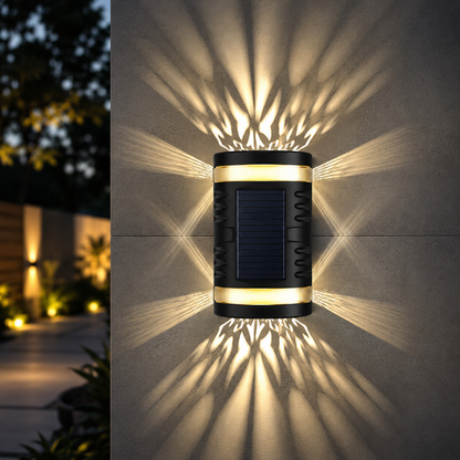 A cylindrical solar wall light mounted on a gray wall at night, casting dramatic starburst light patterns onto the wall. The fixture has a black body with a dark blue solar panel in the center and warm yellow light emitting from the top and bottom bands.