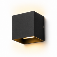 A modern square wall sconce light fixture with a matte black exterior and a warm gold interior, shown illuminated against a plain white background.