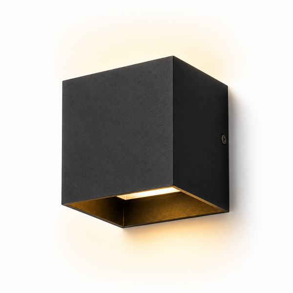 A modern square wall sconce light fixture with a matte black exterior and a warm gold interior, shown illuminated against a plain white background.