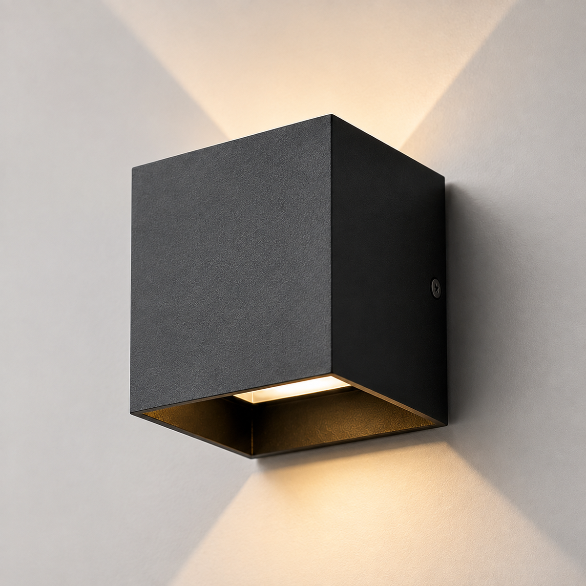 A modern black square wall sconce with a textured exterior and a warm light glowing from the open bottom, mounted on a light gray wall.