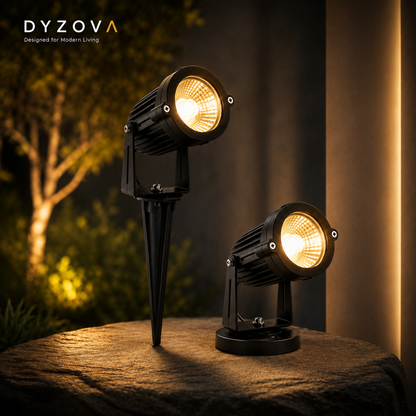 Two black DYZOVA outdoor landscape spotlights with stake and base options, shown lit on a stone surface with a warm garden scene in the background.