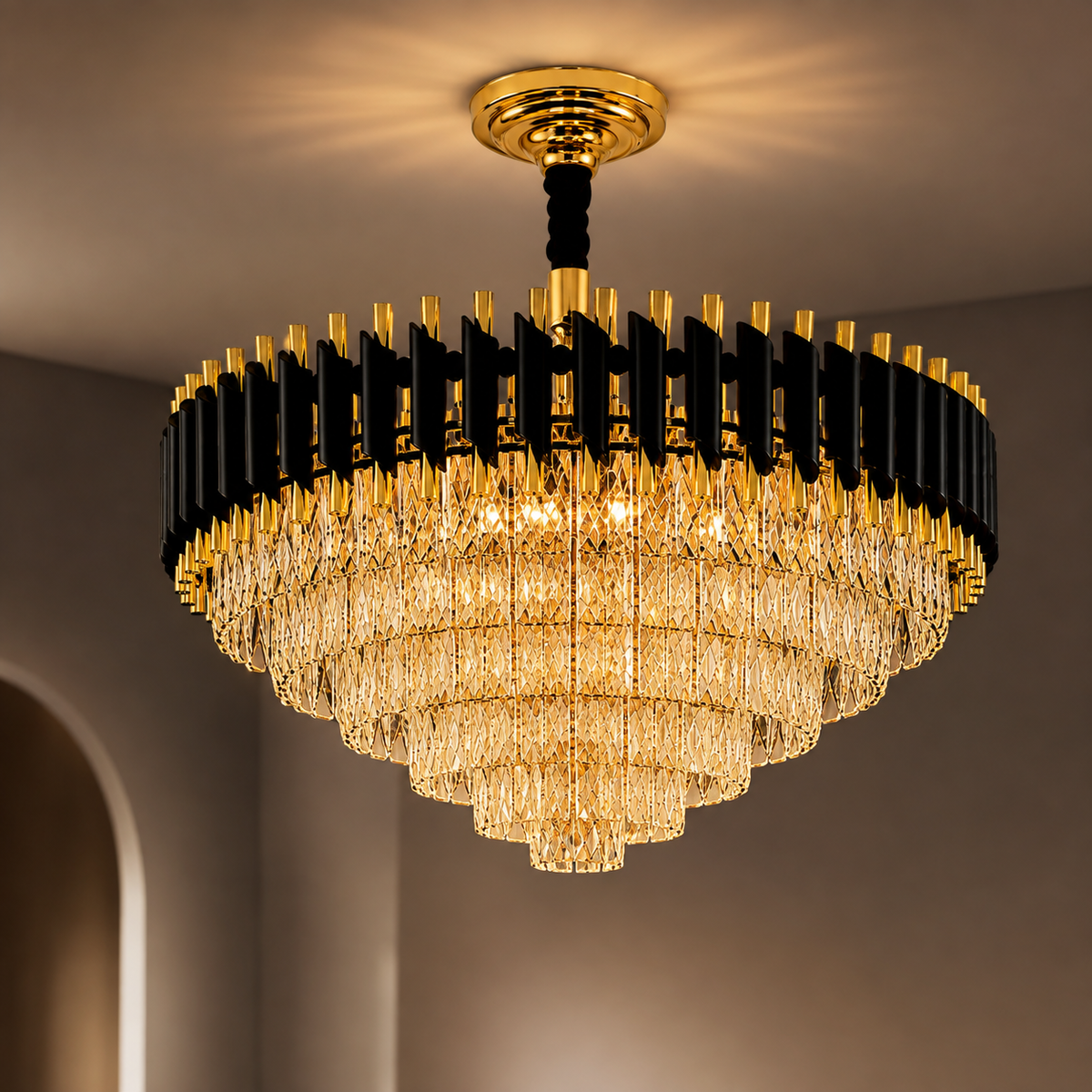 A large modern chandelier with a gold and black metal frame and cascading clear crystal prisms, shown illuminated in a warm light against a neutral interior wall.