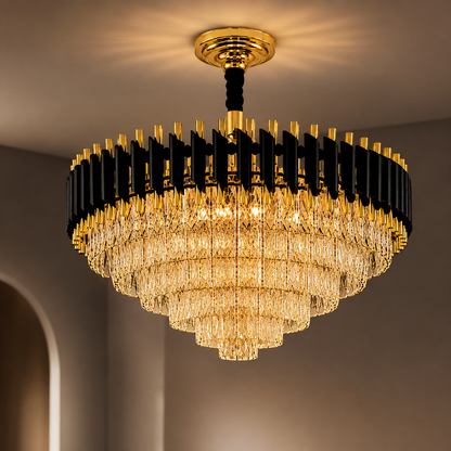 A large modern chandelier with a gold and black metal frame and cascading clear crystal prisms, shown illuminated in a warm light against a neutral interior wall.